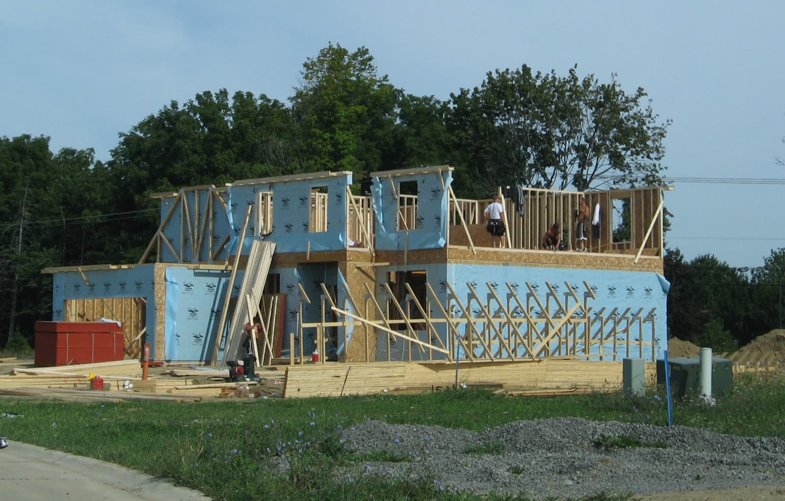 A two-story house under construction with wood framing and sheathing, workers on the upper floor